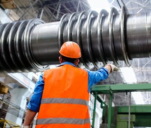An engineer wearing an orange hard hat and an orange vest investigates a manufacturing part in a warehouse.