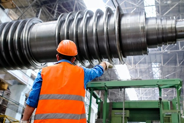 An engineer wearing an orange hard hat and an orange vest investigates a manufacturing part in a warehouse.